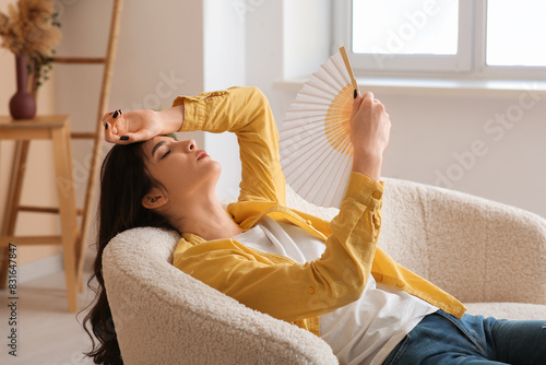 Schilderij op canvas Young woman with hand fan suffering from heat on white sofa in living room