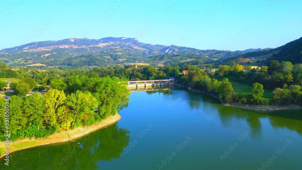 Enchanting aerial pullback shot from Lago di San Ruffino, ranging from the lake shores on the left with a blue pathway and a white small boat, to the green trees surrounding the bridge in the middle Enchanting aerial pullback shot from Lago di San Ruffino, ranging from the lake shores on the left with a blue pathway and a white small boat, to the green trees surrounding the bridge in the middle