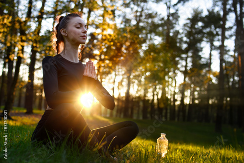 A young beautiful woman in a tight-fitting tracksuit sits on the grass in the woods in lotus pose and meditates.