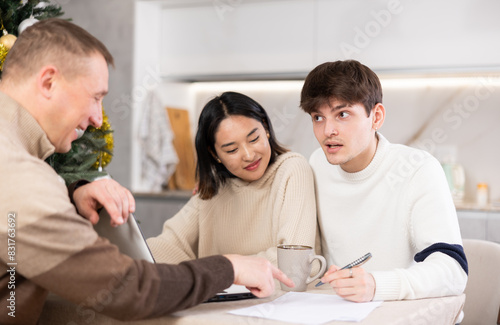 Male realtor discusses the terms of contract with couple in office of real estate agency  offering various accommodation options against backdrop of a Christmas tree