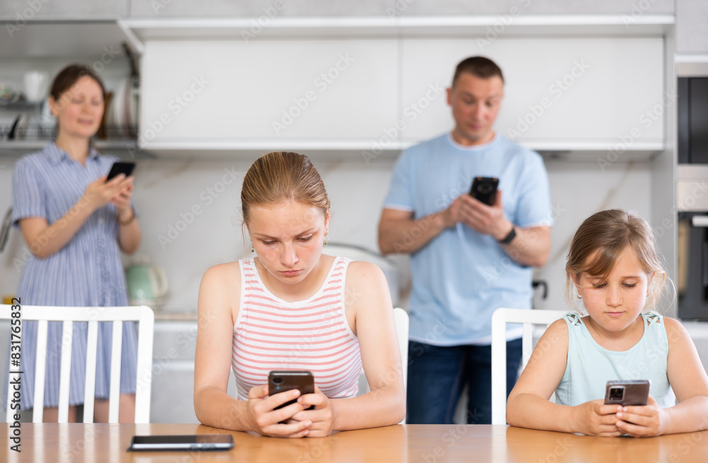 Fototapeta premium Sister girls sitting near kitchen table with phones in hands and flipping through tape and liking photos of friends, parents surf Internet in background