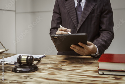 A businessman in a formal suit signs a contract, hands close up, suggesting possible bribery, at a wooden desk with legal documents, scales, and a wooden gavel representing legislation. photo