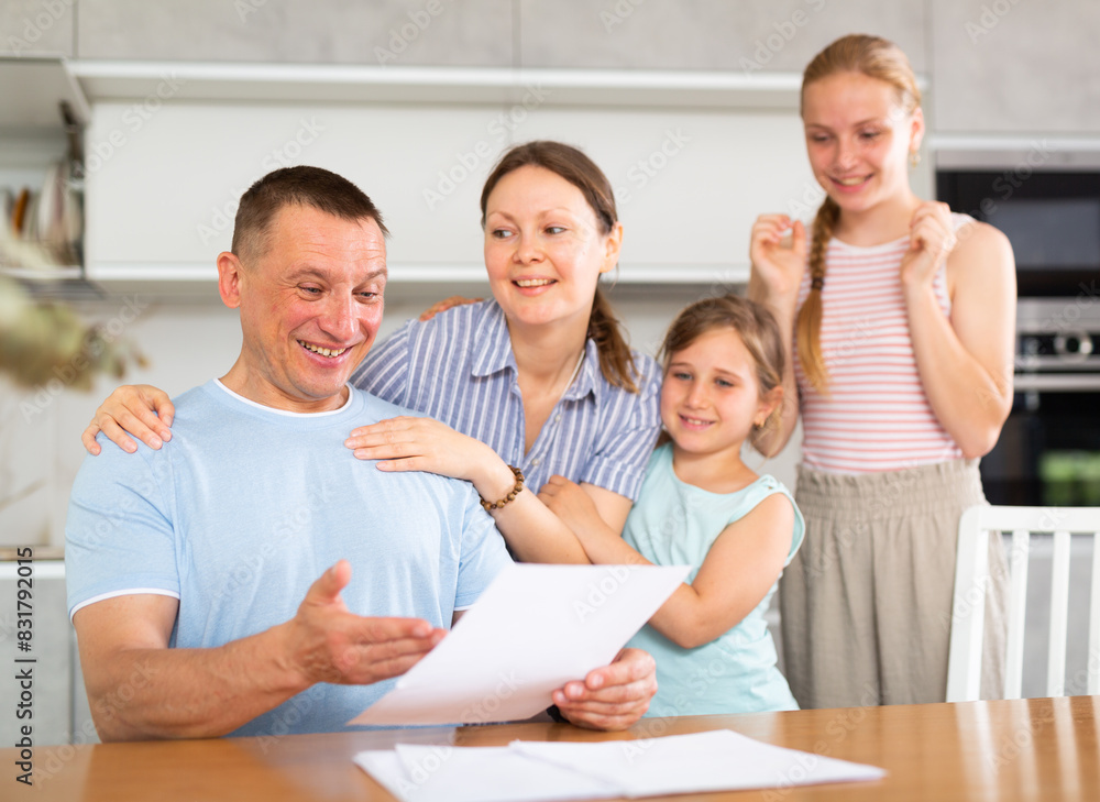 Fototapeta premium Happy family receiving written notification of positive decision from bank or credit organization about approval of loan or mortgage. Cheerful man with wife and teenage daughters reading some papers