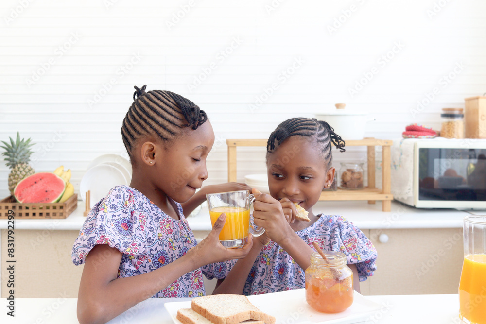 African twin girl sister with curly hair braid African hairstyle eat bread toast with jam and drink orange juice at kitchen. Happy smiling kid sibling have breakfast together. Cute children in family. African twin girl sister with curly hair braid African hairstyle eat bread toast with jam and drink orange juice at kitchen. Happy smiling kid sibling have breakfast together. Cute children in family.