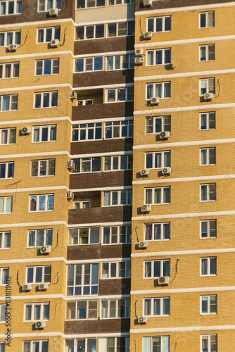 A section of a multi-storey apartment block with a patterned facade with numerous windows and balconies. The exterior cladding of the building is yellow in colour with flecks of brown.