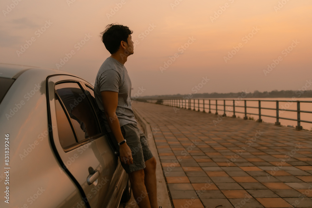 Fototapeta premium Asian man standing relaxing and leaning the car look at nature at sunset time, looking out the river, happy travel concept.