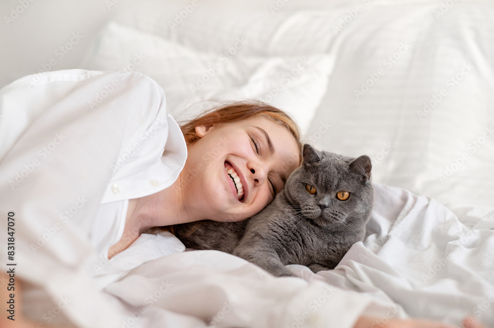 Domestic pet and child friendship and love, smiling teenage girl laying on bed, resting together with gray cat, white neutral bedroom Domestic pet and child friendship and love, smiling teenage girl laying on bed, resting together with gray cat, white neutral bedroom