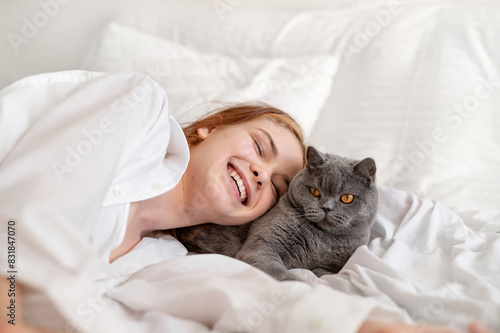 Domestic pet and child friendship and love, smiling teenage girl laying on bed, resting together with gray cat, white neutral bedroom