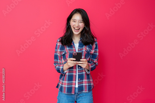 Excited Young Woman in Plaid Shirt Holding Smartphone and Smiling Broadly on Pink Background - Generative AI © RojOct