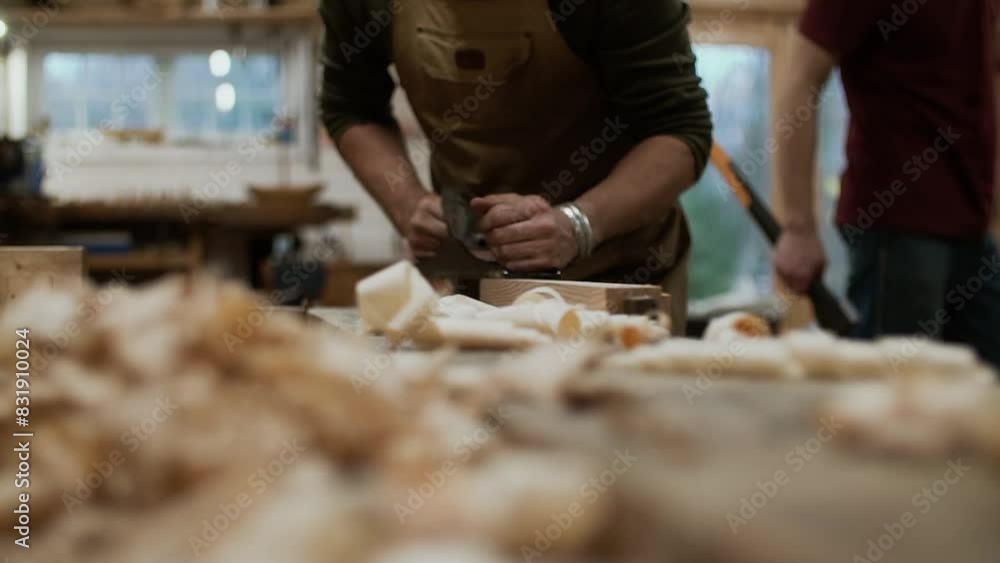 Closeup with a man working with handheld plan in slow motion shaping the wood. Wood shavings drop in front of the camera. Man with an axe in the background. Closeup with a man working with handheld plan in slow motion shaping the wood. Wood shavings drop in front of the camera. Man with an axe in the background.