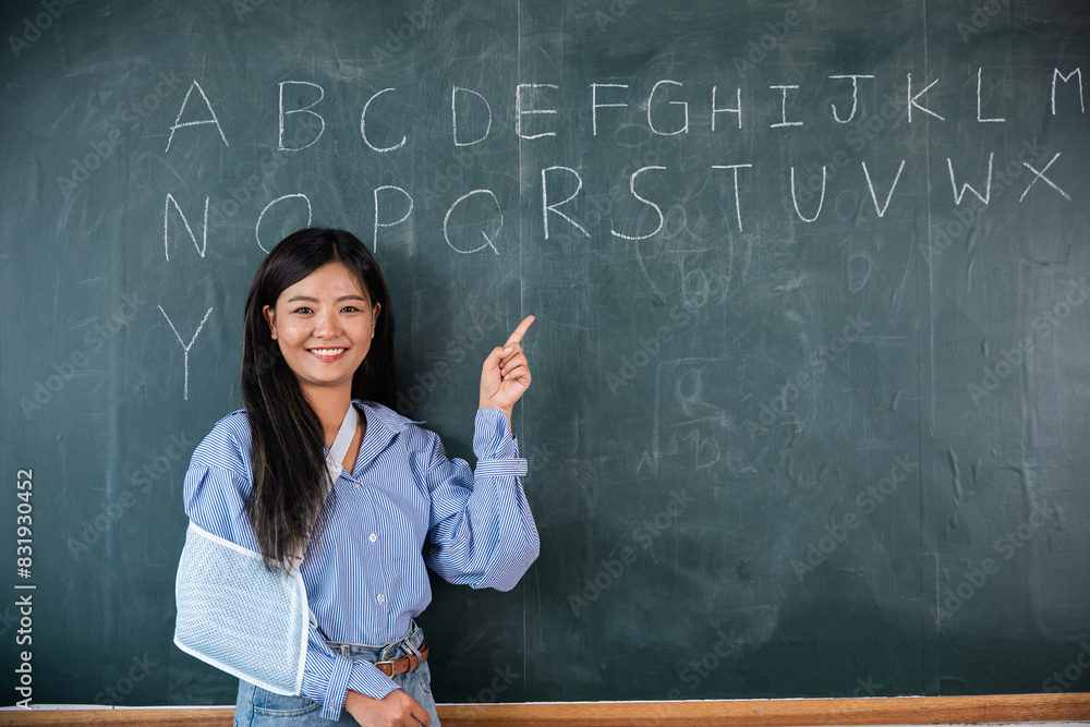 A woman is pointing at the alphabet on a blackboard. Arm broken. Teacher confident broken arm after accident and wear arm splint for treatment, sling support hand A woman is pointing at the alphabet on a blackboard. Arm broken. Teacher confident broken arm after accident and wear arm splint for treatment, sling support hand