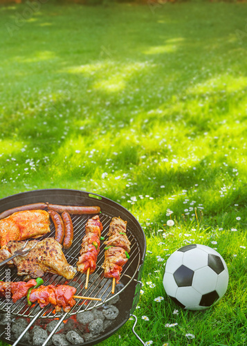  Delicious grilled meat with vegetables sizzling over the coals on barbecue with a soccer ball on the green meadow in the background. Poster. photo
