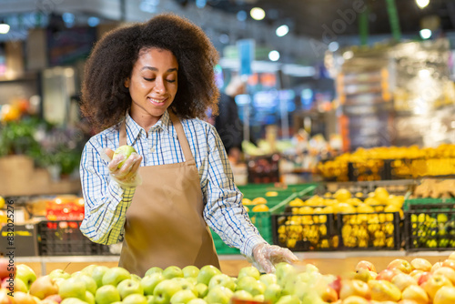 Smiling woman in apron sorting fresh apples at a grocery store, showcasing a vibrant produce section photo