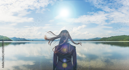 風に髪をなびかせ自然あふれる湖畔の岸に佇むスーツ姿の女性の後ろ姿　夏の太陽の強い日差しとレンズフレア　青空と雲を反射する湖面　遠くの山々に海岸　アウトドア・バカンス・夏休み・キャンプ・旅行・観光・リラクゼーションのイメージ photo