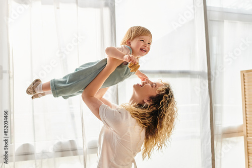 Fotografie A curly mother joyfully lifts her toddler daughter up into the air, expressing love and playfulness at home