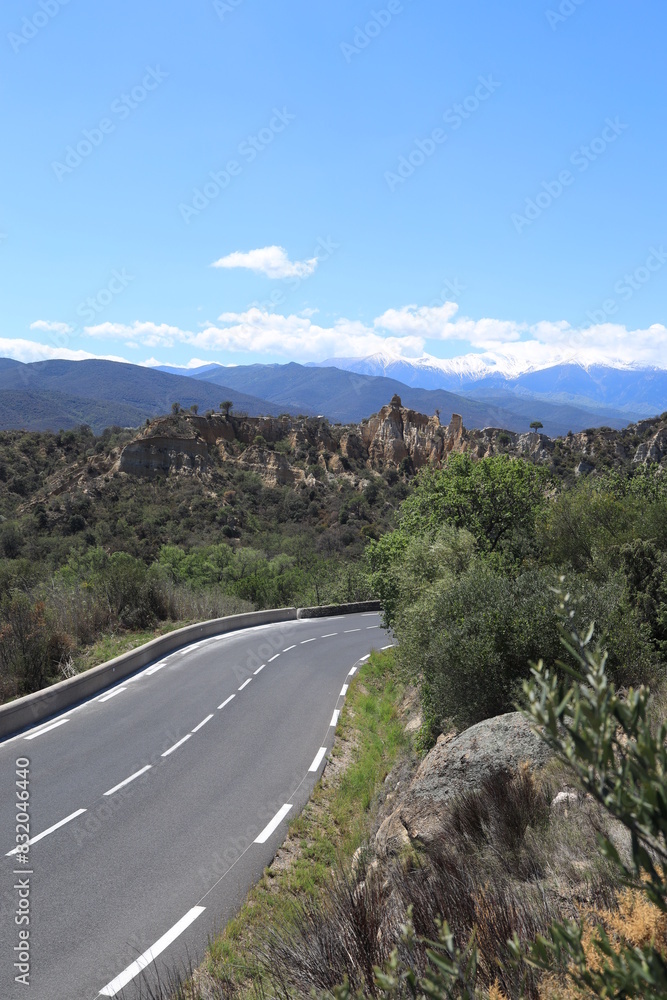 Landscape near Ille-sur-Têt in Pyrénées-Orientales department, southern France featuring winding road leading towards rock chimney formations with Canigou mountain visible in background Landscape near Ille-sur-Têt in Pyrénées-Orientales department, southern France featuring winding road leading towards rock chimney formations with Canigou mountain visible in background
