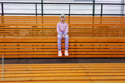 A girl sits on a wooden bench in a sports tribune with her hands on her knees. Child athlete in a purple sports suit and hat. Parallel lines on background with copy space.