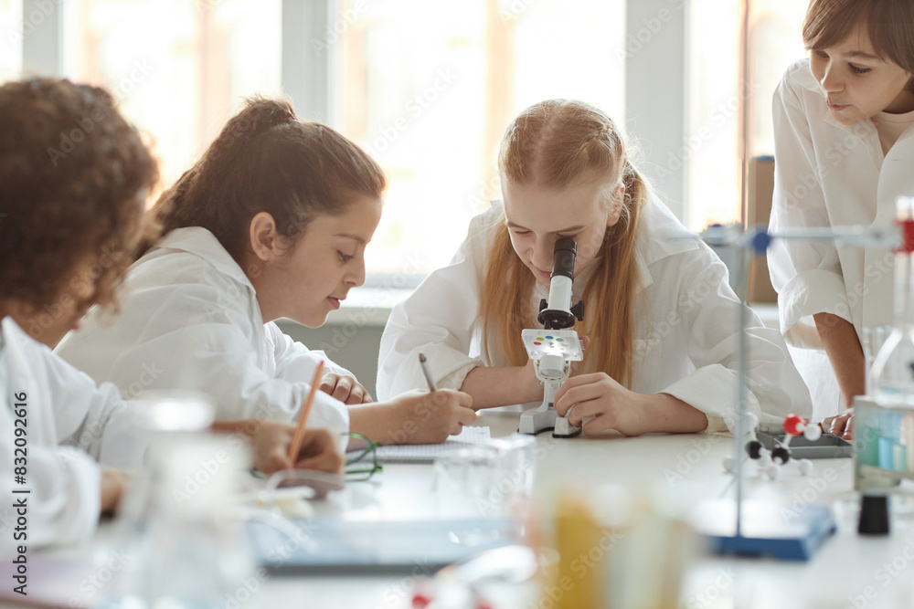 Multi-ethnic group of middle school students wearing lab coats doing experiments with use of microscope during Chemistry lesson Multi-ethnic group of middle school students wearing lab coats doing experiments with use of microscope during Chemistry lesson