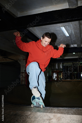 Vertical flash long shot of joyful gen Z guy wearing red hoodie and blue jeans riding skateboard in skatepark, copy space