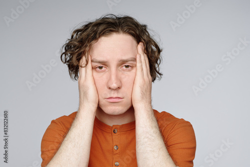 Medium closeup studio portrait of young tired man with curly hair posing for camera with hands covering his ears photo