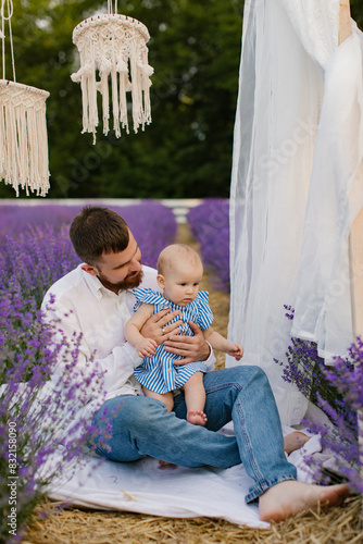 Bearded man in white shirt is sitting on a white blanket and playing with his little daughter in a lavender field. Dad with a baby. Lifestyle. 