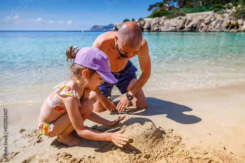 Little girl and her father playing with sand enjoying their time together, on a beautiful beach with clear blue waters and a scenic backdrop