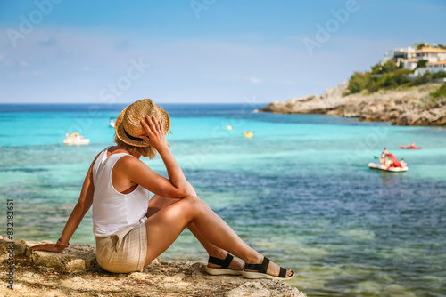 A woman in a straw hat, sitting by the beach, looking out at the clear blue sea, highlighting a serene Mallorca travel moment
