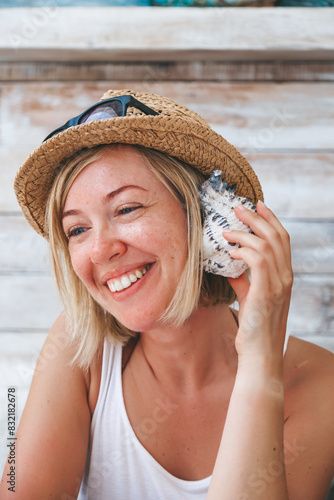 A smiling happy woman wearing a straw hat with sunglasses, holding a seashell close to her ear, seemingly hearing its whisper