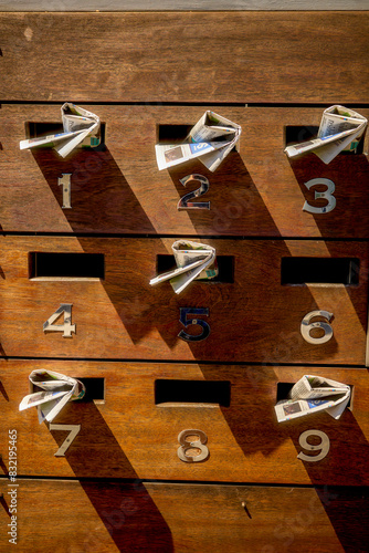 A vertical array of wooden mailboxes with numbers from 1 to 9, each containing rolled-up newspapers.