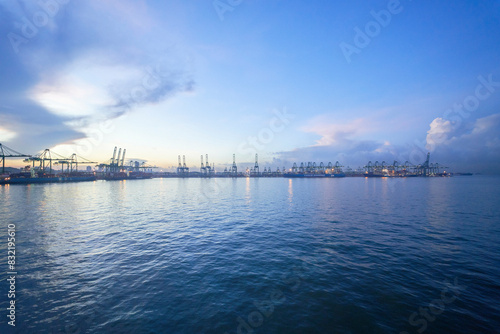 Dawn breaks over a busy port with rows of cranes lining the docks, and container handling operations underway, against a backdrop of a calm sea and a clear sky with soft clouds. photo