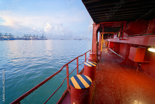 The image captures a vibrant view from the deck of a ship showing red and yellow railings, calm sea water, and container cranes at a port in the distance under a partly cloudy sky. photo