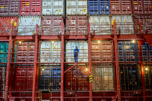 A worker stands on a metal stairway amidst a large stack of colorful shipping containers at night, illuminated by ambient lights.