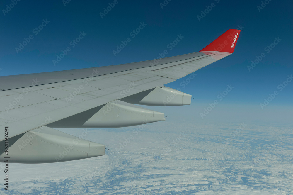 An airplane wing with a red tip soars above the clouds and ice-covered landscape below, capturing the majesty of flight in a clear blue sky. An airplane wing with a red tip soars above the clouds and ice-covered landscape below, capturing the majesty of flight in a clear blue sky.
