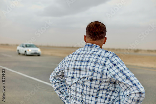 A man is seen from behind watching a car drive away on a deserted road under a cloudy sky.