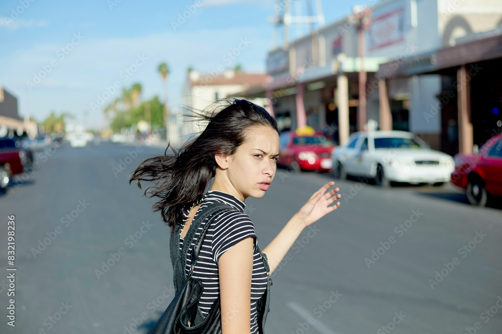 A woman is walking on a sunny street with her hair blowing in the wind; shops and parked cars line the road behind her. A woman is walking on a sunny street with her hair blowing in the wind; shops and parked cars line the road behind her.