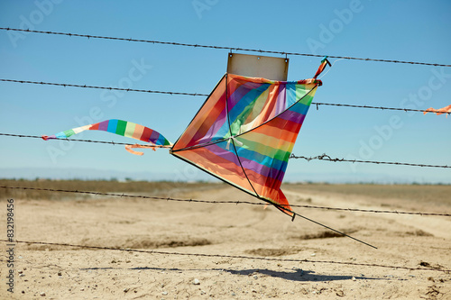 A colorful kite is entangled in a barbed wire fence against a clear blue sky in a barren landscape. photo