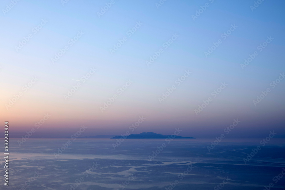 A serene view of a tranquil sea at dusk with a silhouetted island in the distance under a gradient sky transitioning from warm pink to cool blue.