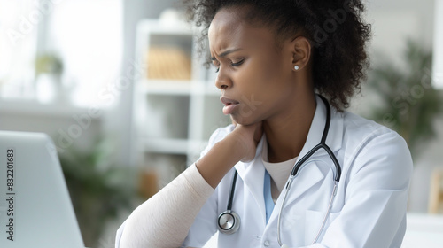 An African American woman doctor in a hospital, holding her neck in discomfort, expressing frustration and the strain of overwork, with a laptop present