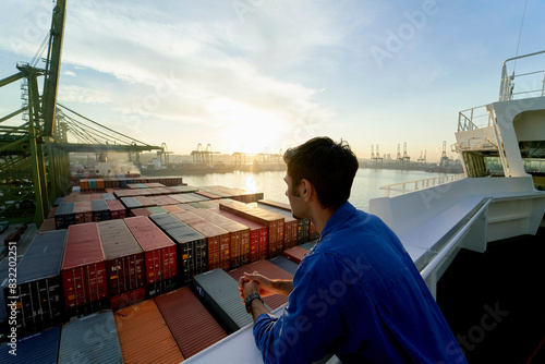 A person stands on a ship, gazing out at a harbor filled with shipping containers during sunrise.