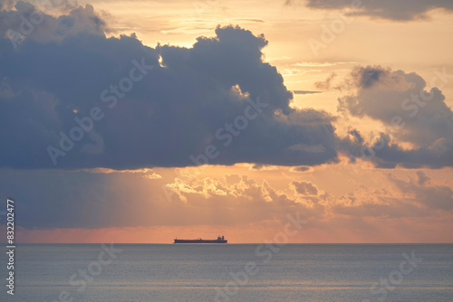 A serene ocean scene at dusk with a cargo ship visible on the horizon, backlit by the soft, warm hues of the setting sun peeking through scattered clouds. photo