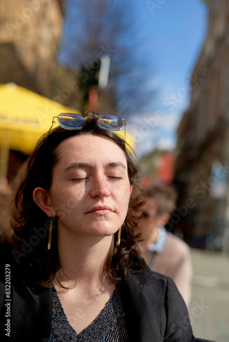 A young adult is enjoying the warmth of the sunlight with their eyes closed and sunglasses on their head, against an urban backdrop. photo