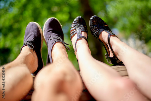 A couple have extended their legs upwards against a backdrop of green tree foliage, showcasing their different styles of footwear under the bright sunlight.