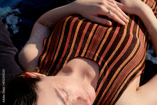 A young woman with freckles and a nose ring lies on a striped cloth, her eyes closed and hands resting on her chest in a relaxed pose.