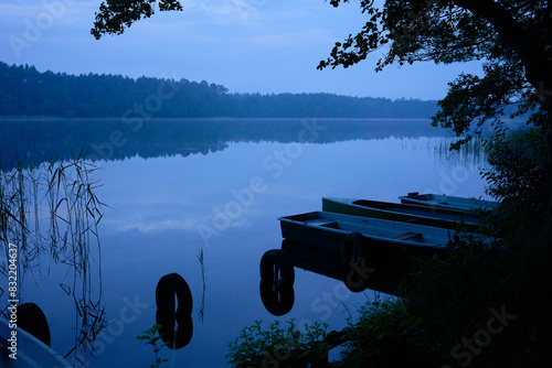 A serene lakeside scene in the early morning featuring rowboats tied to a dock, with a calm reflective water surface and a hint of mist hovering above the lake. photo