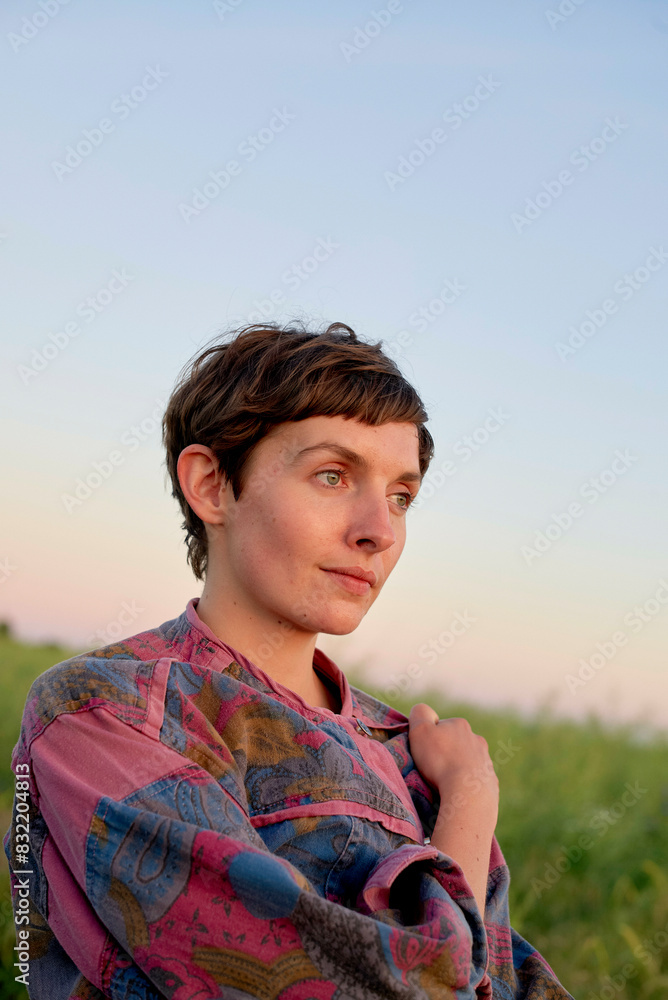 A person with short hair is wearing a colorful printed shirt and gazing to the side with a soft expression against a blurred natural background during what appears to be dusk. A person with short hair is wearing a colorful printed shirt and gazing to the side with a soft expression against a blurred natural background during what appears to be dusk.