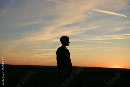 Silhouette of an individual standing in a field at sunset, with the sky showing warm colors and streaks of clouds. photo