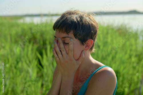 A young woman is sitting outdoors with their eyes covered by their hands, seemingly in a moment of distress or contemplation, with a water body and green reeds in the background.