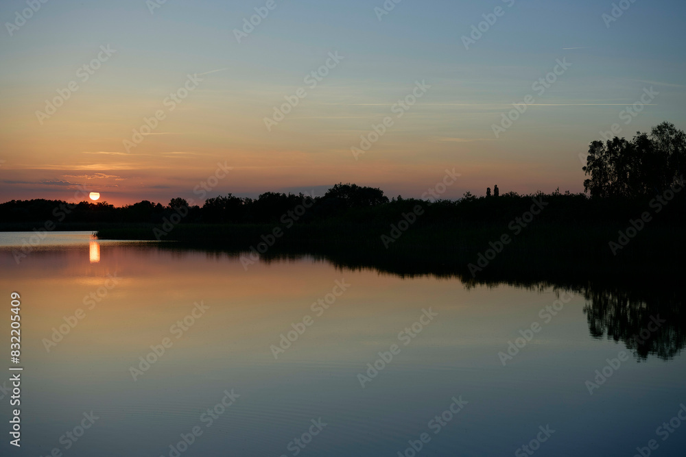 A serene sunset scene over a calm lake with reflections of the diminishing sun and silhouetted landscape against a gradient evening sky. A serene sunset scene over a calm lake with reflections of the diminishing sun and silhouetted landscape against a gradient evening sky.