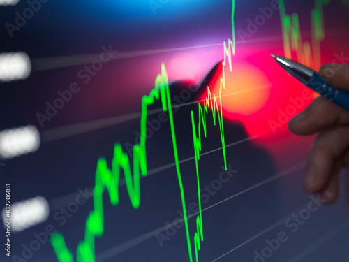 A hand holding a pen points to a bright stock market chart on a digital screen with fluctuating green and red graph lines indicating financial data. photo