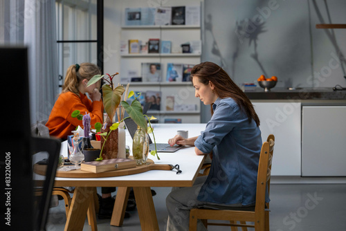 Two women are focused on their respective tasks at a dining table in a well-lit, modern kitchen.